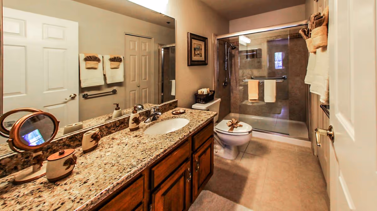 A clean and well-lit bathroom featuring a granite countertop with a sink, a large mirror, wooden cabinets, a toilet, and a glass-enclosed shower with beige tiles. Towels are neatly hung on racks and folded on the toilet seat, and decorative items are placed on the countertop.