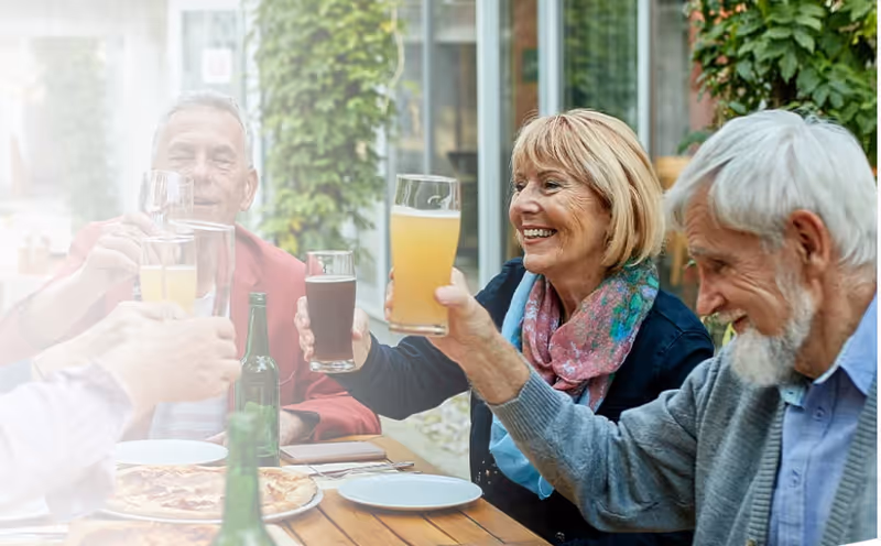 A group of elderly people sitting around a wooden table outdoors, smiling and raising their glasses in a toast. There are plates and a pizza on the table, with greenery and glass windows in the background.