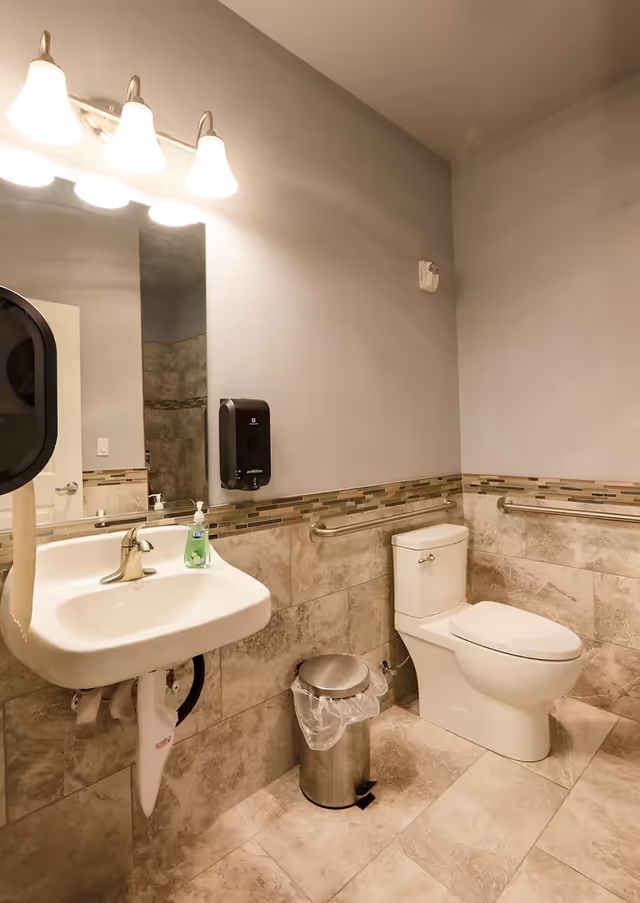 A clean and accessible bathroom with a white toilet, a wall-mounted sink with a soap dispenser, a trash can with a plastic liner, grab bars on the walls, beige tiled floor and walls, and a large mirror above the sink illuminated by three light fixtures.