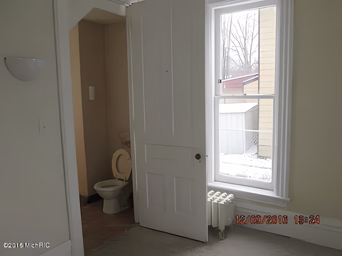 View of a partially open white door leading to a small bathroom with a toilet. Next to the door is a window showing an outdoor scene with snow and a shed. A radiator is positioned below the window.