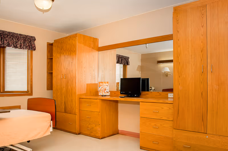 A senior living facility bedroom featuring a single bed with a light-colored bedspread, wooden built-in wardrobes and drawers, a large mirror above the desk area, a small television on the desk, and a window with blinds and a floral valance.