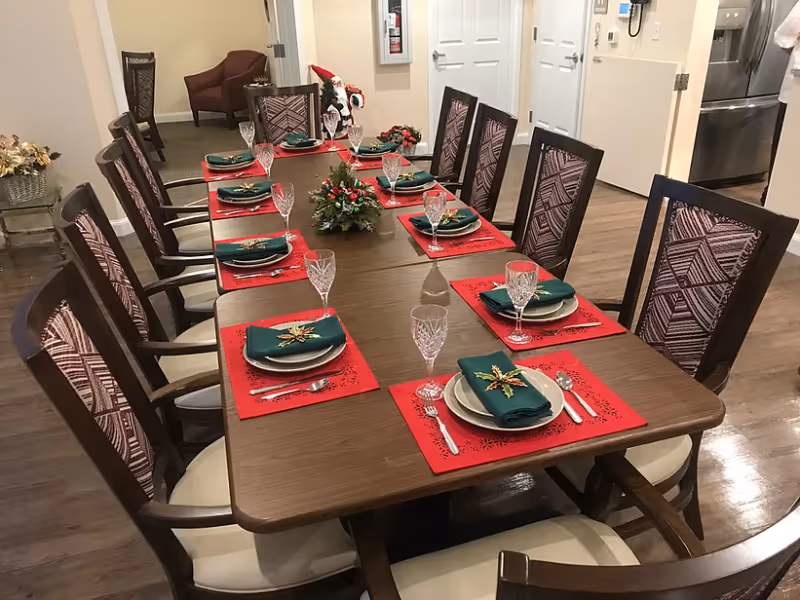 A long wooden dining table set for eight people with red placemats, green napkins decorated with gold stars, white plates, silverware, and crystal glasses. The room has wooden flooring and upholstered chairs with patterned backs. In the background, there is a small seating area with a brown armchair and a stainless steel refrigerator visible through an open doorway.
