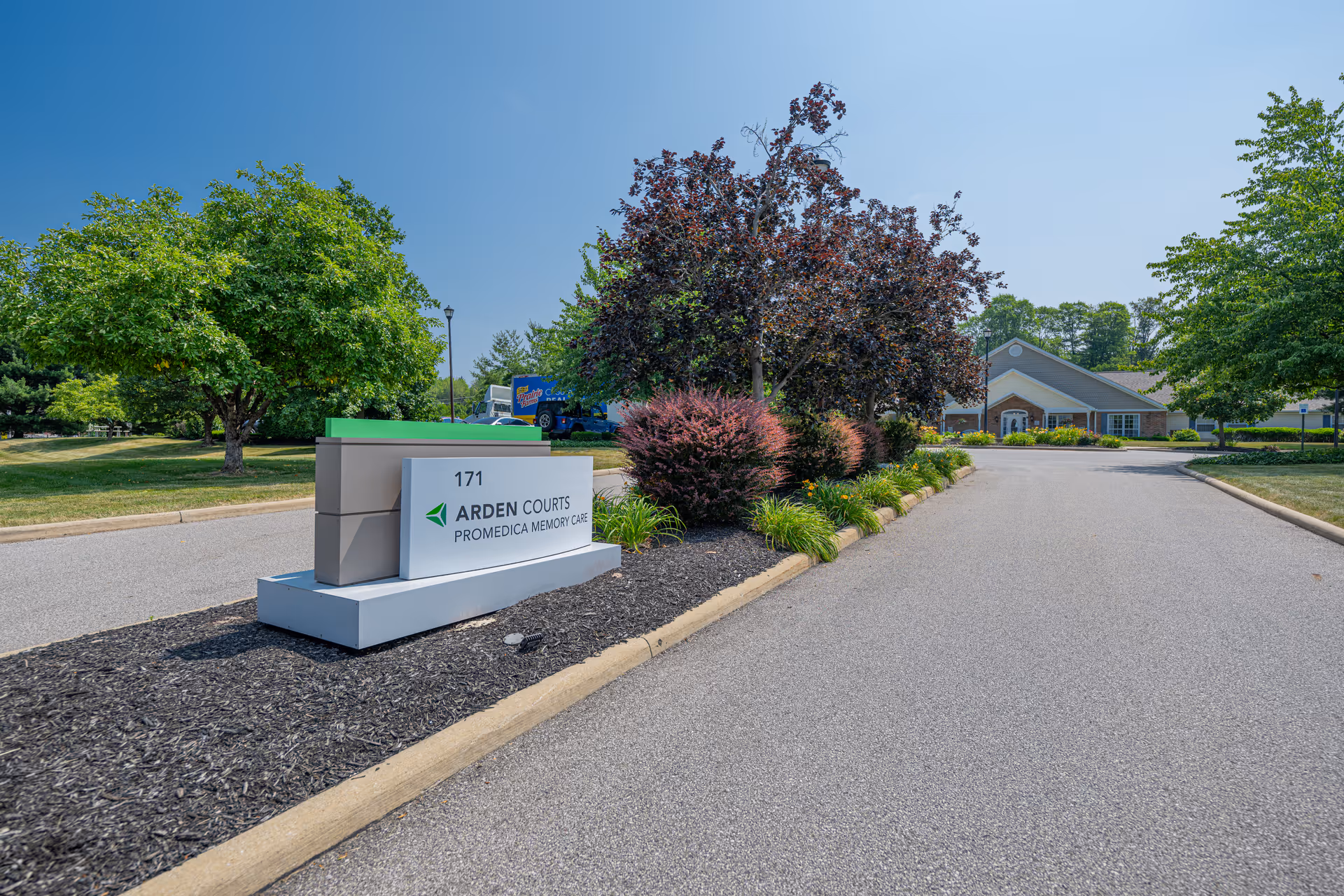 Entrance drive with a landscaped sign that reads 'Arden Courts ProMedica Memory Care' and the facility building visible in the background.