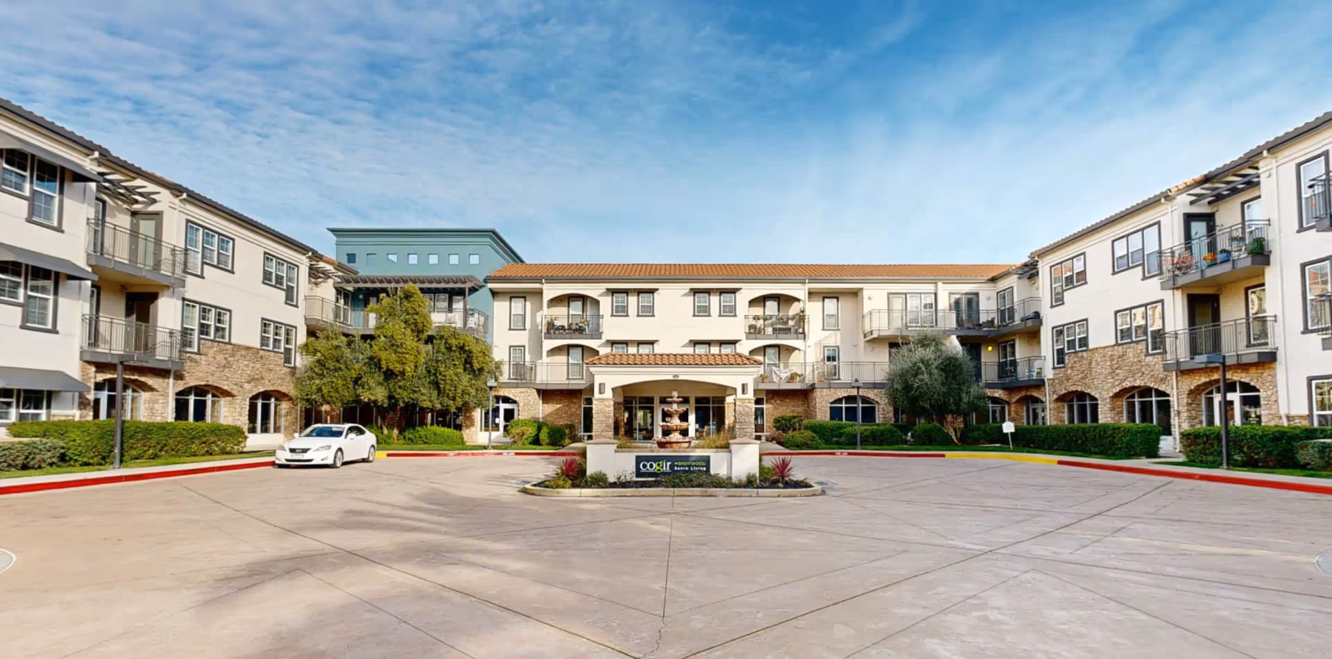 Front exterior view of Cogir of Brentwood senior living facility showing a large driveway with a fountain centerpiece, surrounded by a three-story building with balconies, windows, and some greenery under a partly cloudy sky.