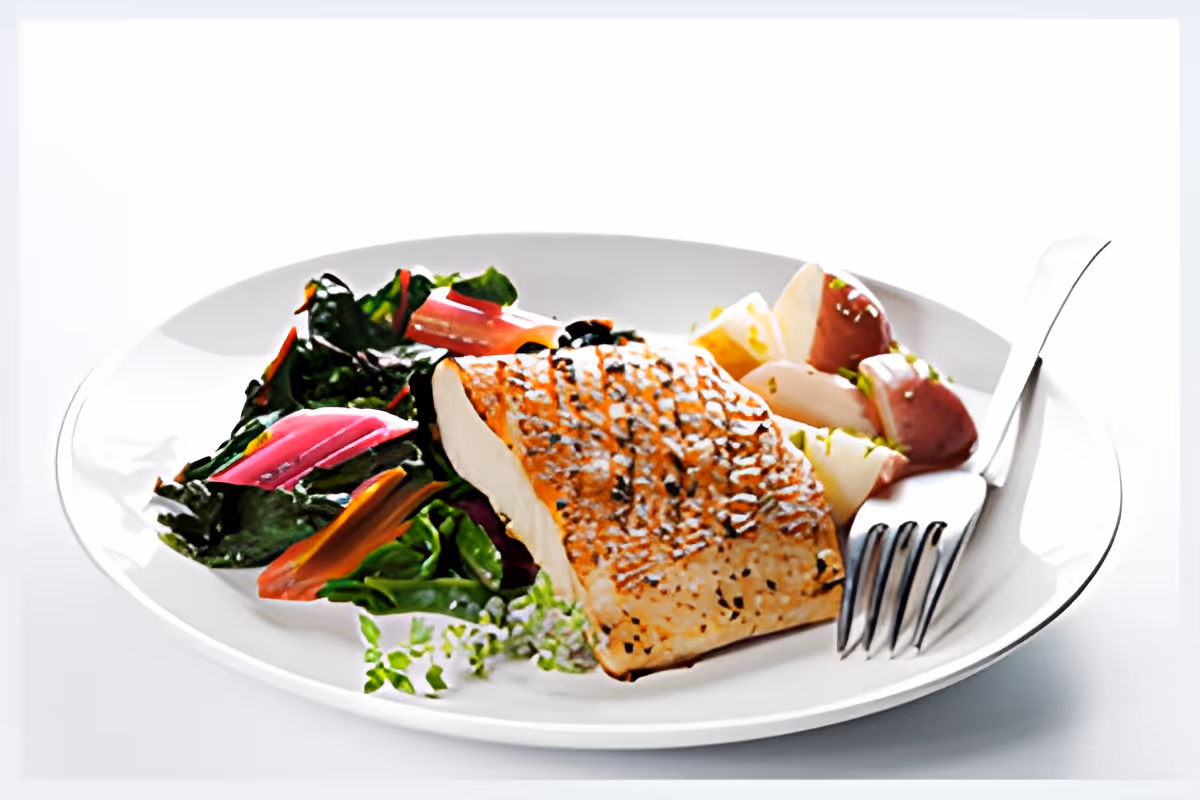 A white plate with a grilled salmon fillet, sautéed leafy greens with colorful stems, and a side of red-skinned potatoes garnished with herbs. A fork and knife are placed on the right side of the plate.