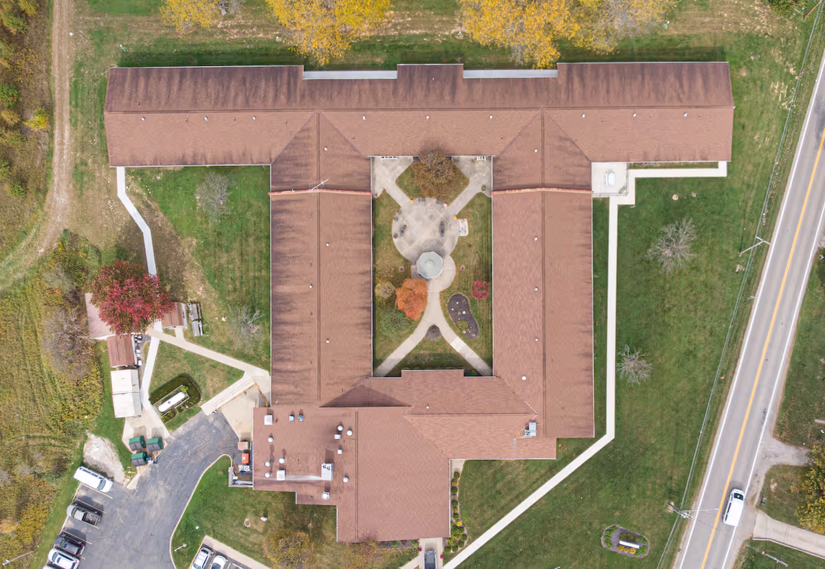 Aerial view of a senior living facility building with a central courtyard garden featuring walkways, trees, and a gazebo. The building has a brown roof and is surrounded by green lawns, a parking area with several cars, and a road on the right side.