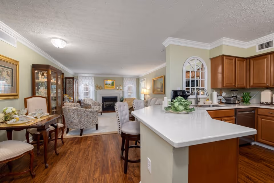 A bright and spacious interior view of a senior living facility showing a kitchen with wooden cabinets and a white countertop island with two cushioned bar stools. Adjacent to the kitchen is a living room area with patterned armchairs, a fireplace, and framed artwork on the walls. The floor is wooden, and the walls are painted light green. A small dining table with chairs is visible on the left side.