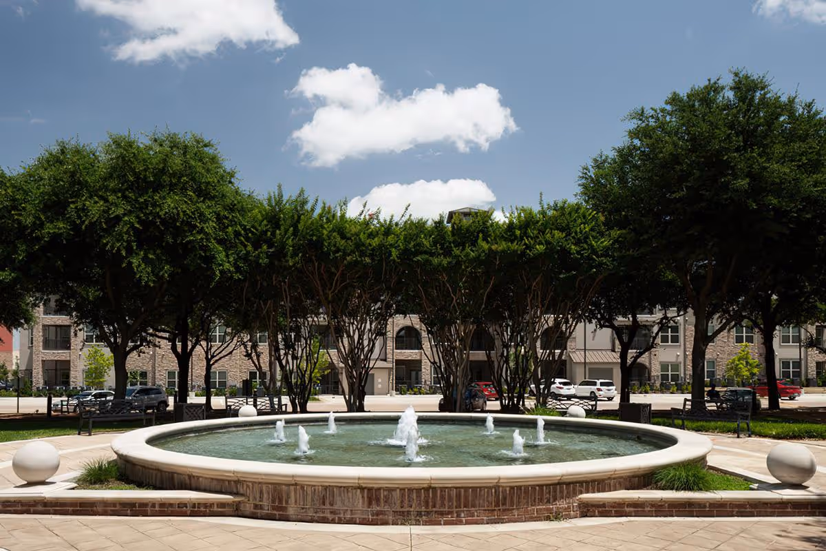 A circular fountain in a paved courtyard with trees and the McDermott Crossing building in the background under a blue sky.
