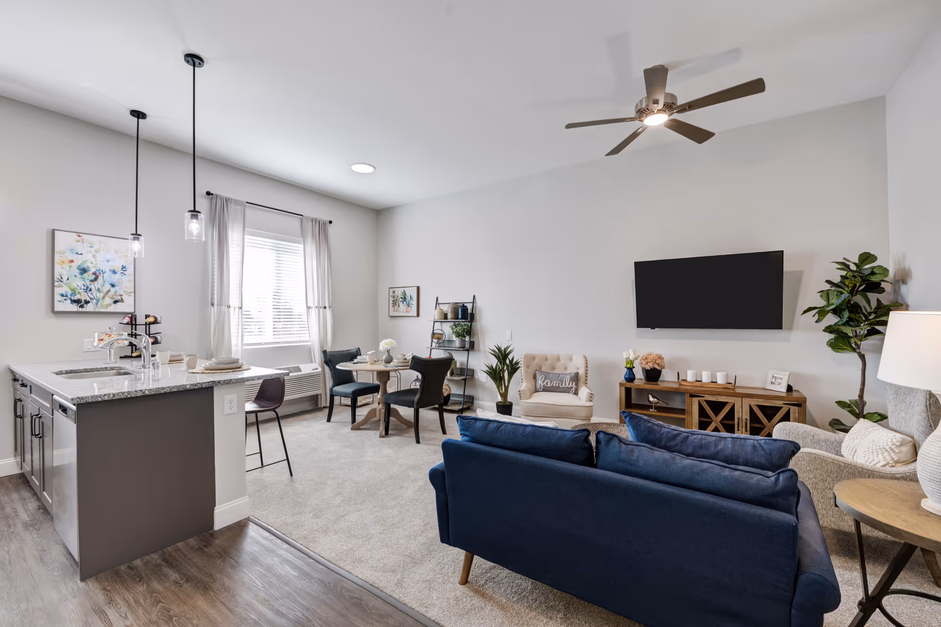 A modern and cozy living room and kitchen area in a senior living facility. The living room features a blue sofa, a beige armchair with a 'family' pillow, a wooden TV stand with a flat-screen TV mounted on the wall, and a tall green plant. The kitchen area has a granite countertop island with two pendant lights hanging above, a sink, and a dishwasher. There is a small dining table with four chairs near a window with light curtains, and the floor transitions from wood in the kitchen to carpet in the living area.