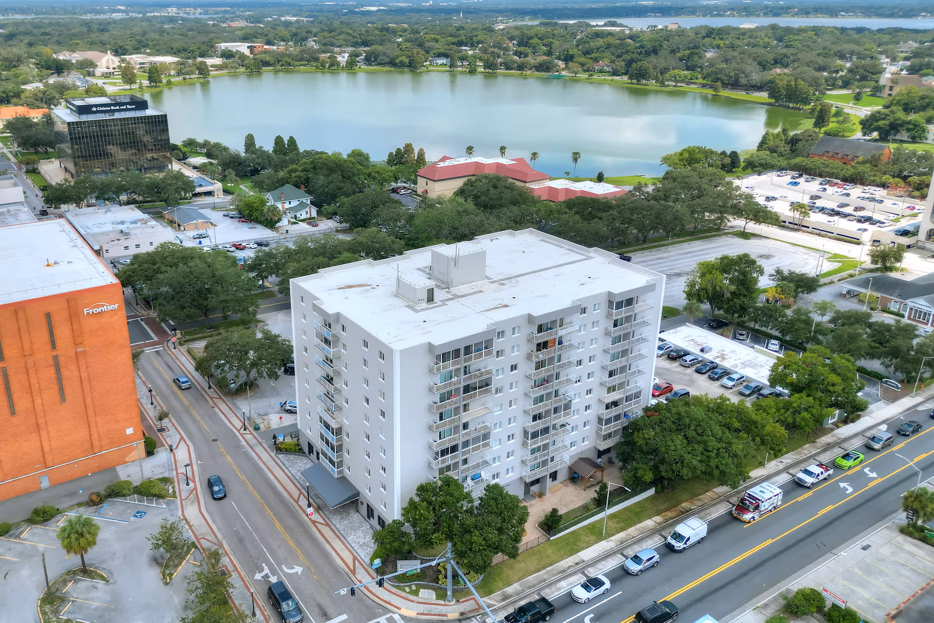 Aerial view of a multi-story residential building named Lake Morton Plaza, surrounded by trees, roads, parked cars, and other buildings, with a large lake and greenery in the background.