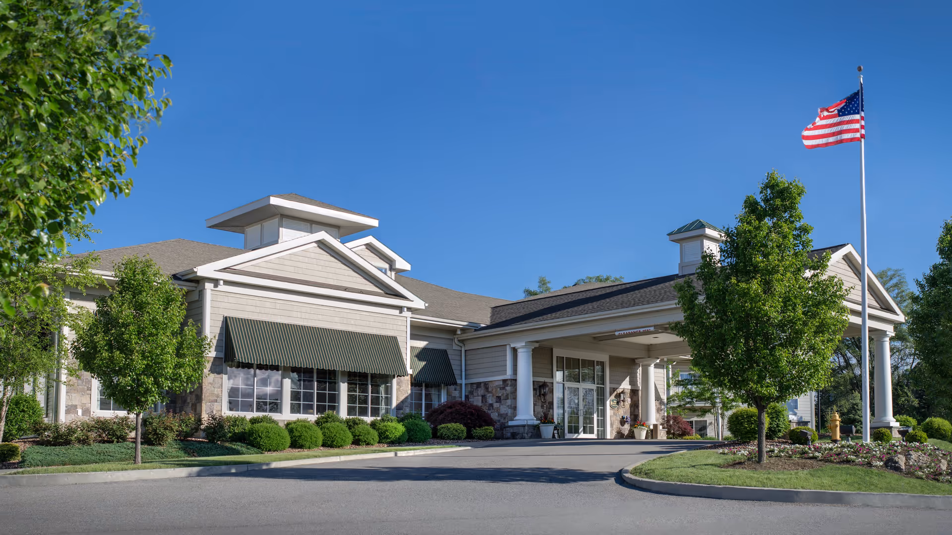 Front entrance of a senior living facility with a covered porte-cochère, landscaped grounds, and an American flag.