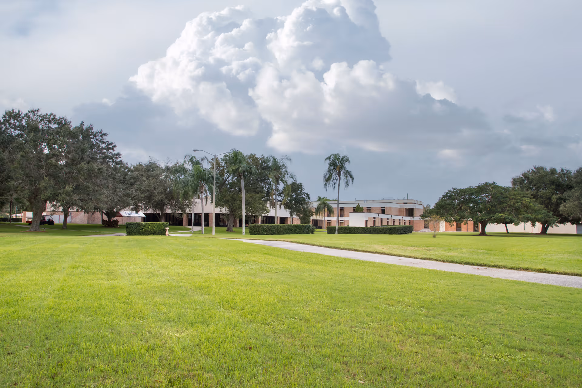 A wide view of a senior living facility with a large green lawn in the foreground, a paved walkway, several trees including palm trees, and a low, light-colored building in the background under a cloudy sky.