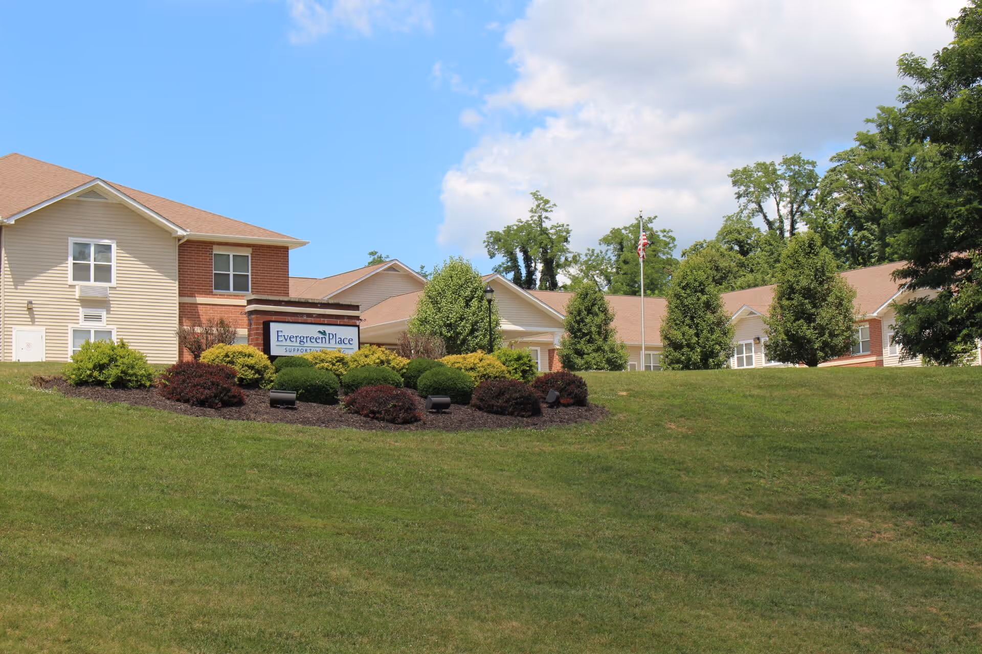 Exterior view of Evergreen Place Supportive Living Alton facility with beige and brick buildings, a manicured lawn, landscaped bushes, and an American flag on a flagpole under a partly cloudy blue sky.