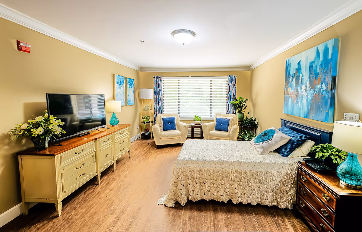 A bright and cozy bedroom in Montclair Assisted Living featuring a bed with a crocheted bedspread, two armchairs with blue cushions near a large window with patterned curtains, a wooden dresser with a flat-screen TV and a vase of flowers, and a nightstand with a turquoise lamp and plants. The walls are painted beige and decorated with blue-themed artwork.