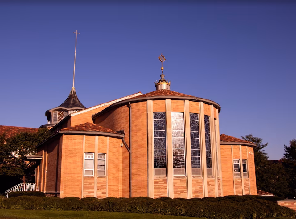 Exterior view of a brick building with large stained glass windows and a tall spire topped with a cross, set against a clear blue sky with green bushes and grass in the foreground.