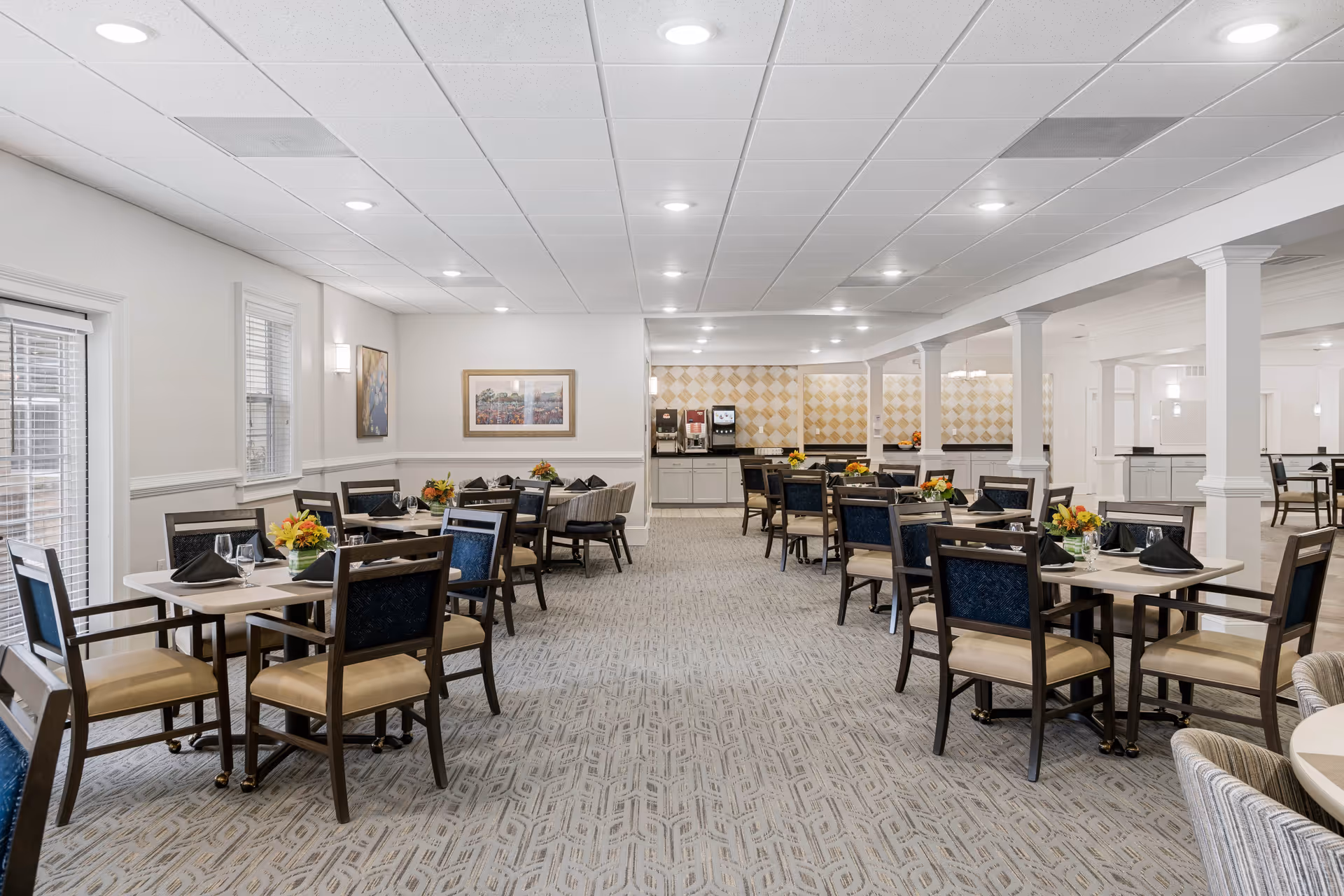 A spacious dining room with multiple tables and chairs arranged neatly. Each table is set with black napkins, glassware, and small floral centerpieces. The room has a light-colored carpet, white walls, and a ceiling with recessed lighting. There is a beverage station with coffee and water dispensers at the far end of the room.