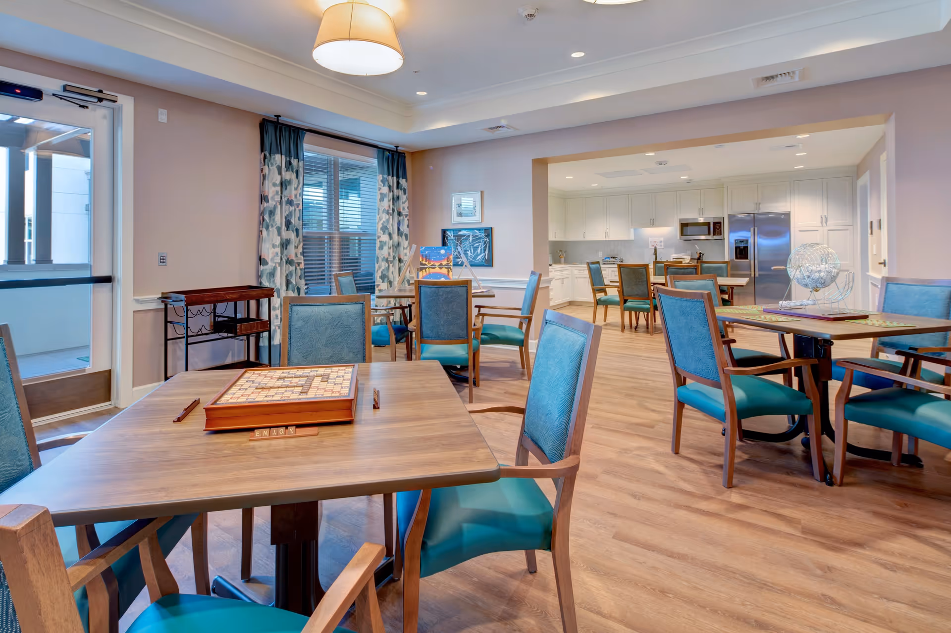 A bright and spacious common area in a senior living facility featuring multiple wooden tables with teal cushioned chairs. The tables have board games and a bingo cage on them. The room has wood flooring, large windows with patterned curtains, and a kitchen area with white cabinets and stainless steel appliances in the background.