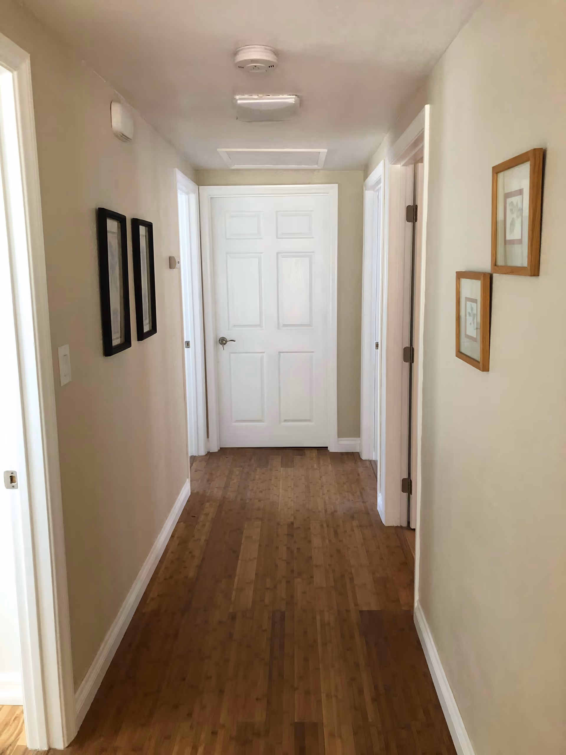 A hallway with wooden flooring and beige walls, featuring four white doors and framed pictures hanging on both walls.
