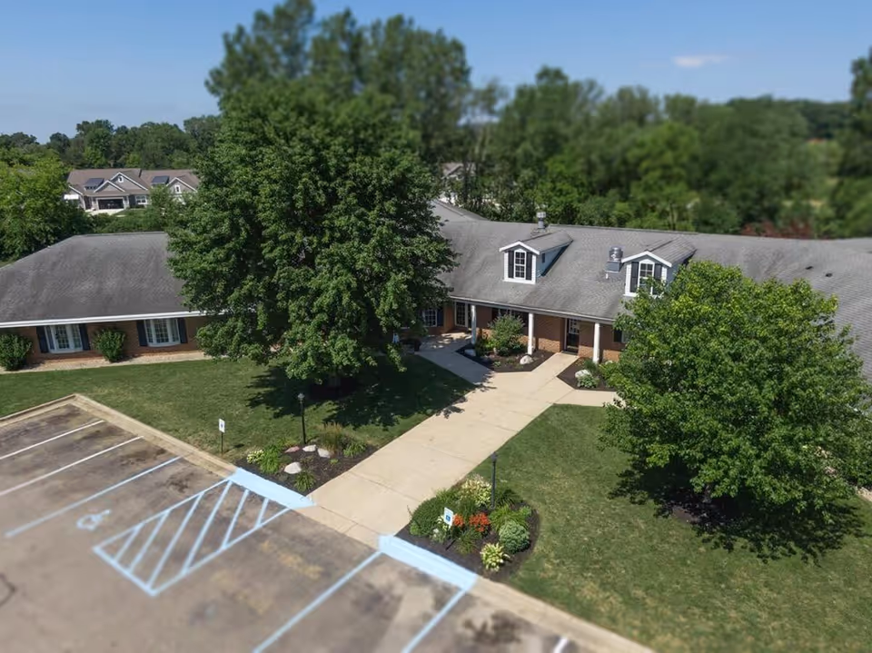 Aerial view of a single-story brick building with a gray shingled roof surrounded by green trees and landscaping. A concrete walkway leads to the entrance, and there is a parking lot with marked spaces, including handicapped spots, in the foreground. The scene is set on a clear, sunny day.