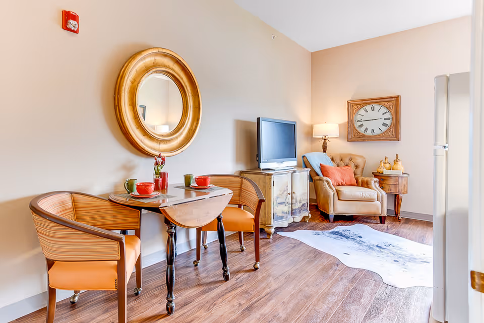 A cozy living area with a small wooden dining table set for two with red and green cups, two wooden chairs with orange cushions, a round gold-framed mirror on the wall, a TV on a decorative cabinet, a beige armchair with a blue and an orange pillow, a side table with decorative items, a large wall clock, a floor lamp, and a white and black patterned rug on wooden flooring.