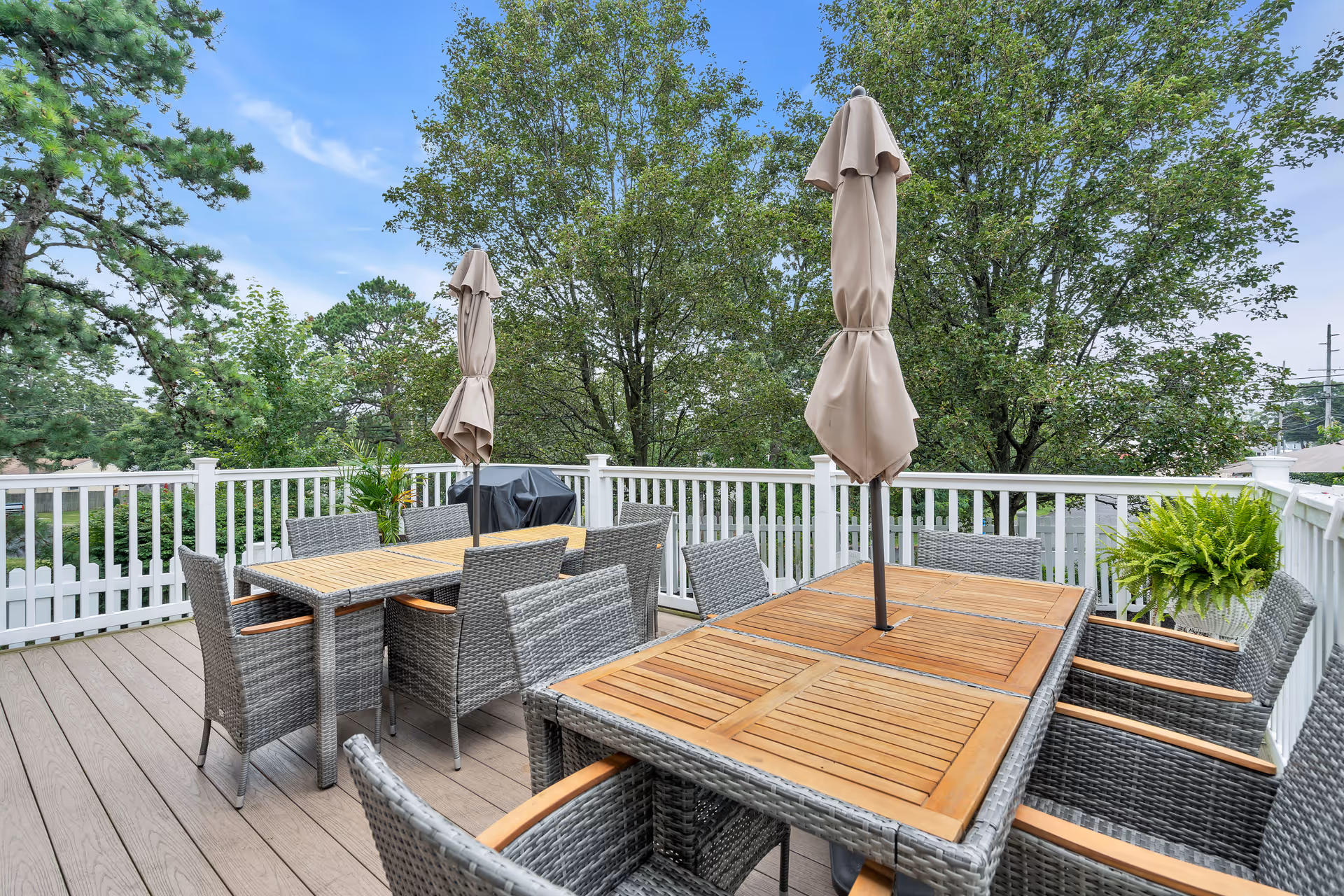 Outdoor patio area with two wooden tables surrounded by wicker chairs with cushions. Each table has a closed beige umbrella in the center. The patio is enclosed by a white railing and surrounded by green trees under a blue sky.