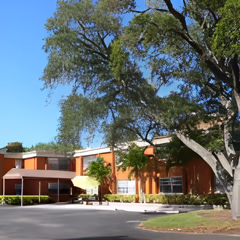Front exterior of a two-story orange senior living building with a large tree and driveway.