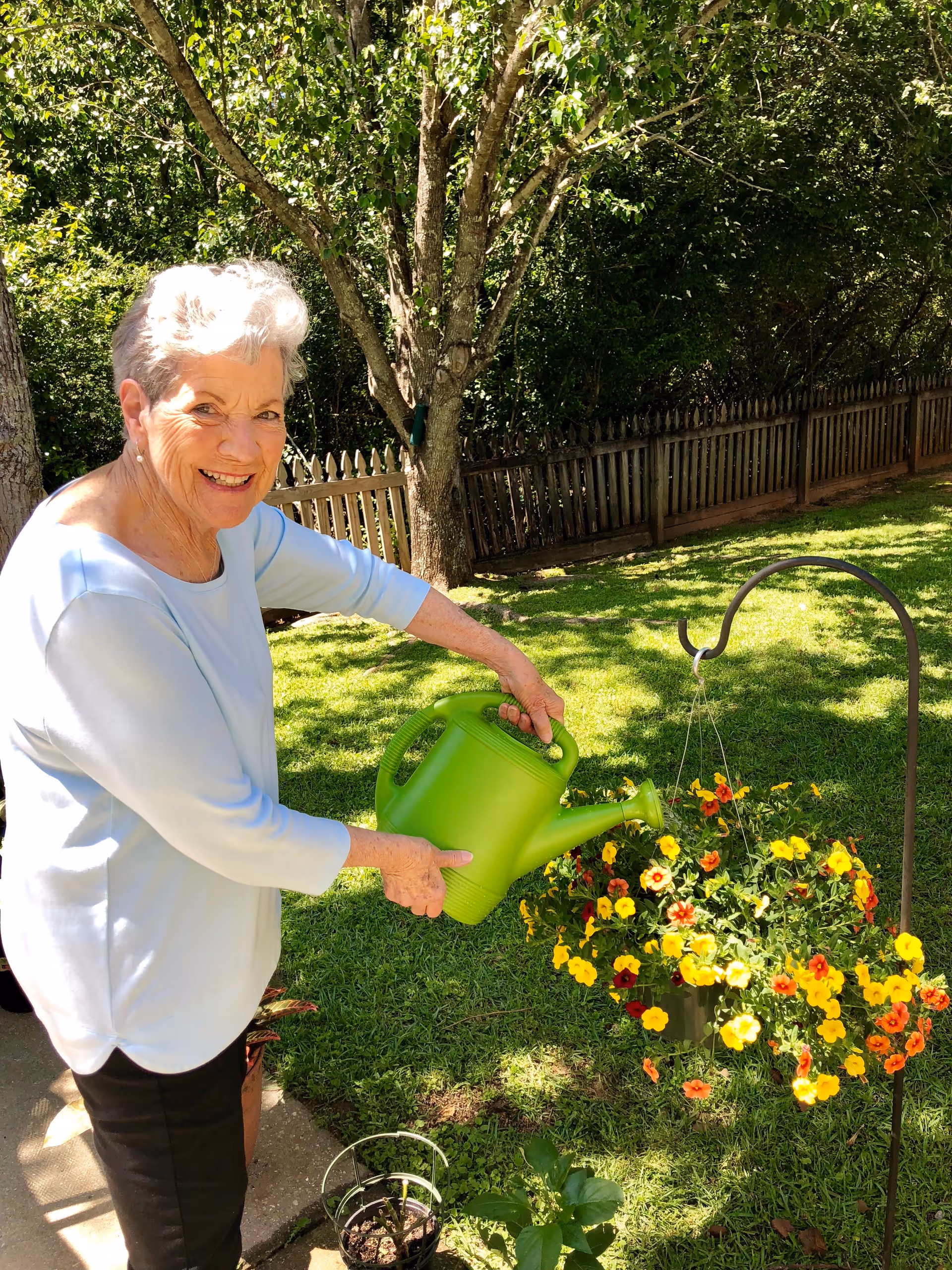 An elderly woman with short white hair wearing a light blue long-sleeve shirt and black pants is smiling while watering a hanging basket of colorful yellow and orange flowers in a sunny garden with green grass, trees, and a wooden fence in the background.