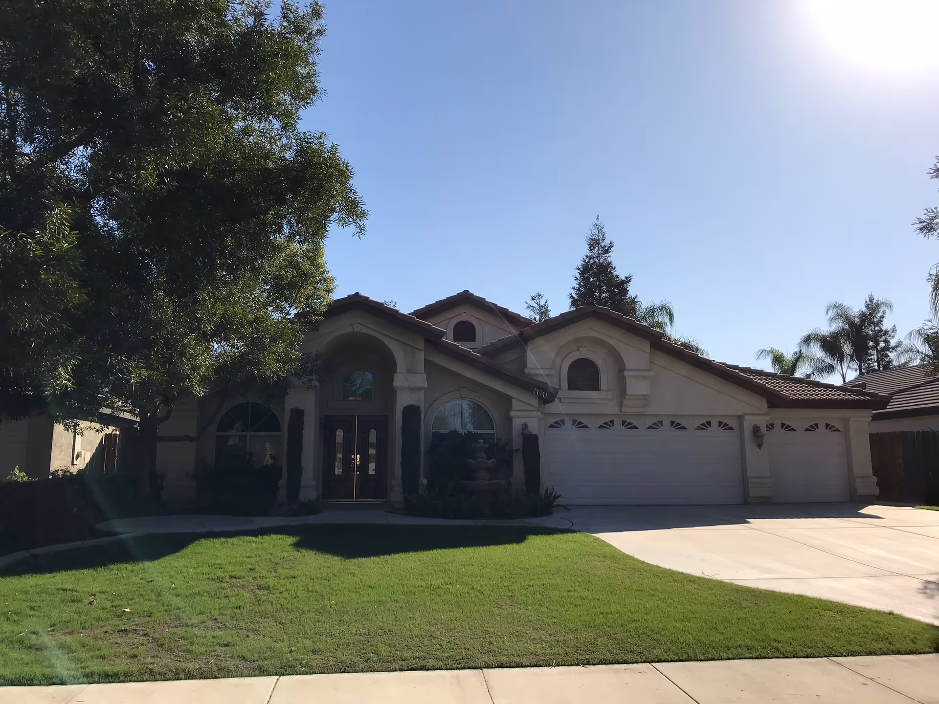Front exterior view of a single-story residential-style building with a three-car garage, arched windows, and a well-maintained green lawn with trees on the left side under a clear blue sky.