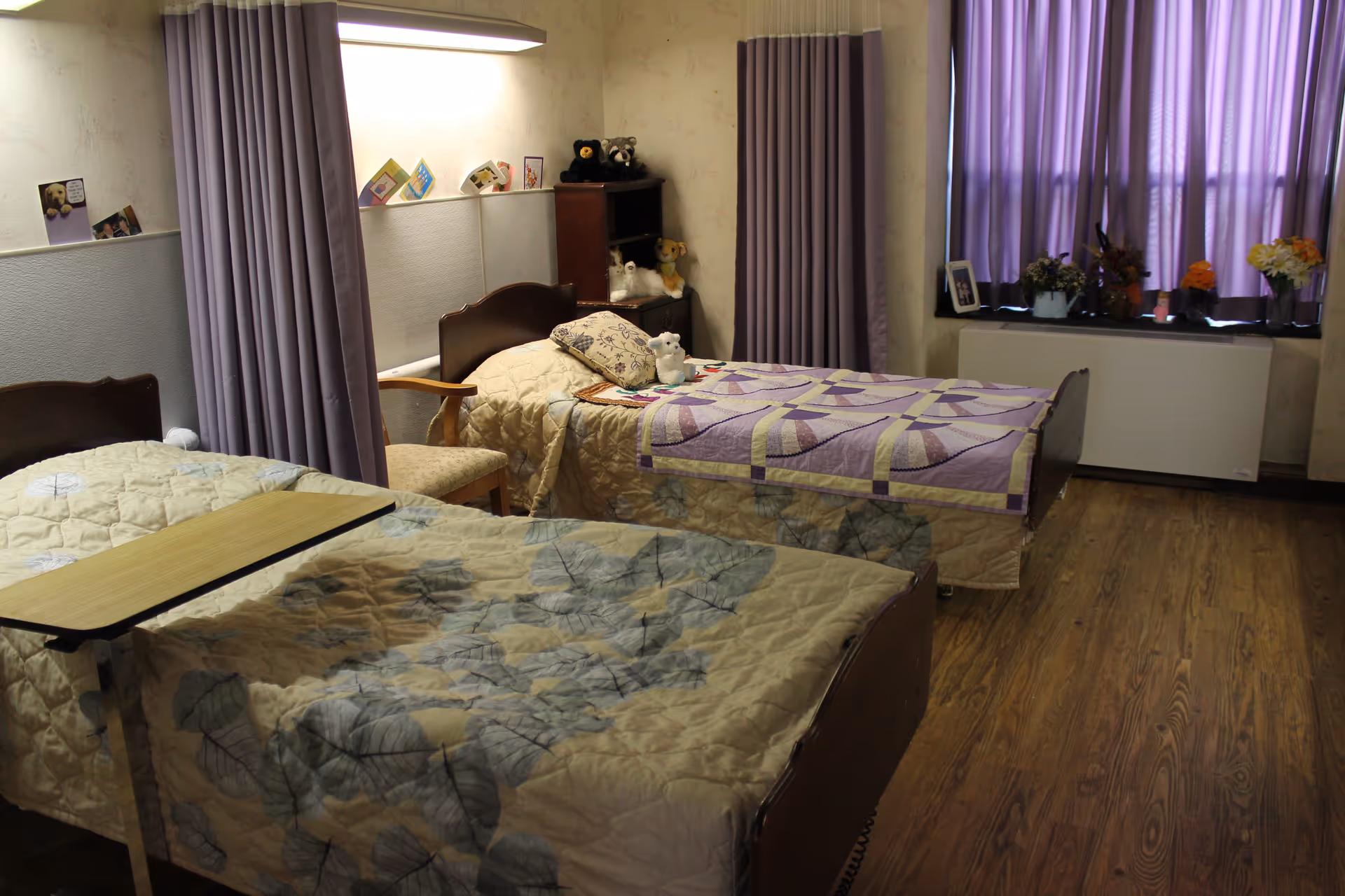 A room with two single beds separated by a curtain. One bed has a beige quilt with a leaf pattern and a wooden overbed table, the other has a purple and beige patchwork quilt with a pillow and a small stuffed animal. There is a wooden chair between the beds, a small shelving unit with stuffed animals and cards, purple curtains covering a window, and a wooden floor.