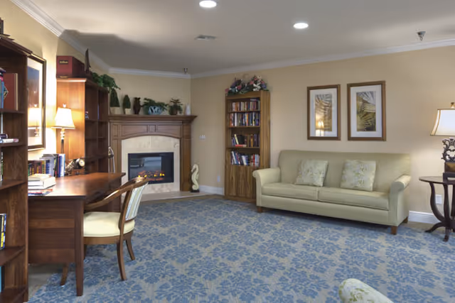 A cozy living room with a beige sofa adorned with patterned cushions, two framed pictures on the wall above it, a wooden bookshelf filled with books, and a fireplace with decorative items on the mantel. There is also a wooden desk with a chair, a table lamp, and a blue patterned carpet covering the floor.