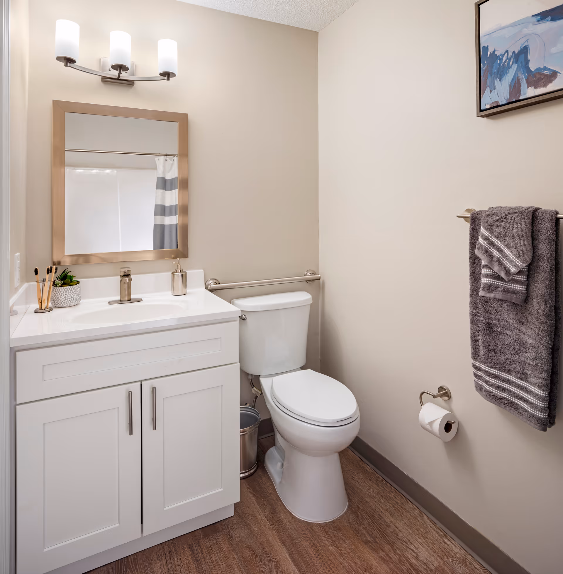 A clean and modern bathroom featuring a white vanity with a sink, a large rectangular mirror with a metallic frame, and a three-light fixture above. Next to the vanity is a white toilet with a metal grab bar behind it. On the right wall, there is a towel rack holding two gray towels and a toilet paper holder with a roll of toilet paper. The floor has wood-like vinyl flooring, and a framed abstract painting hangs on the wall above the towel rack.