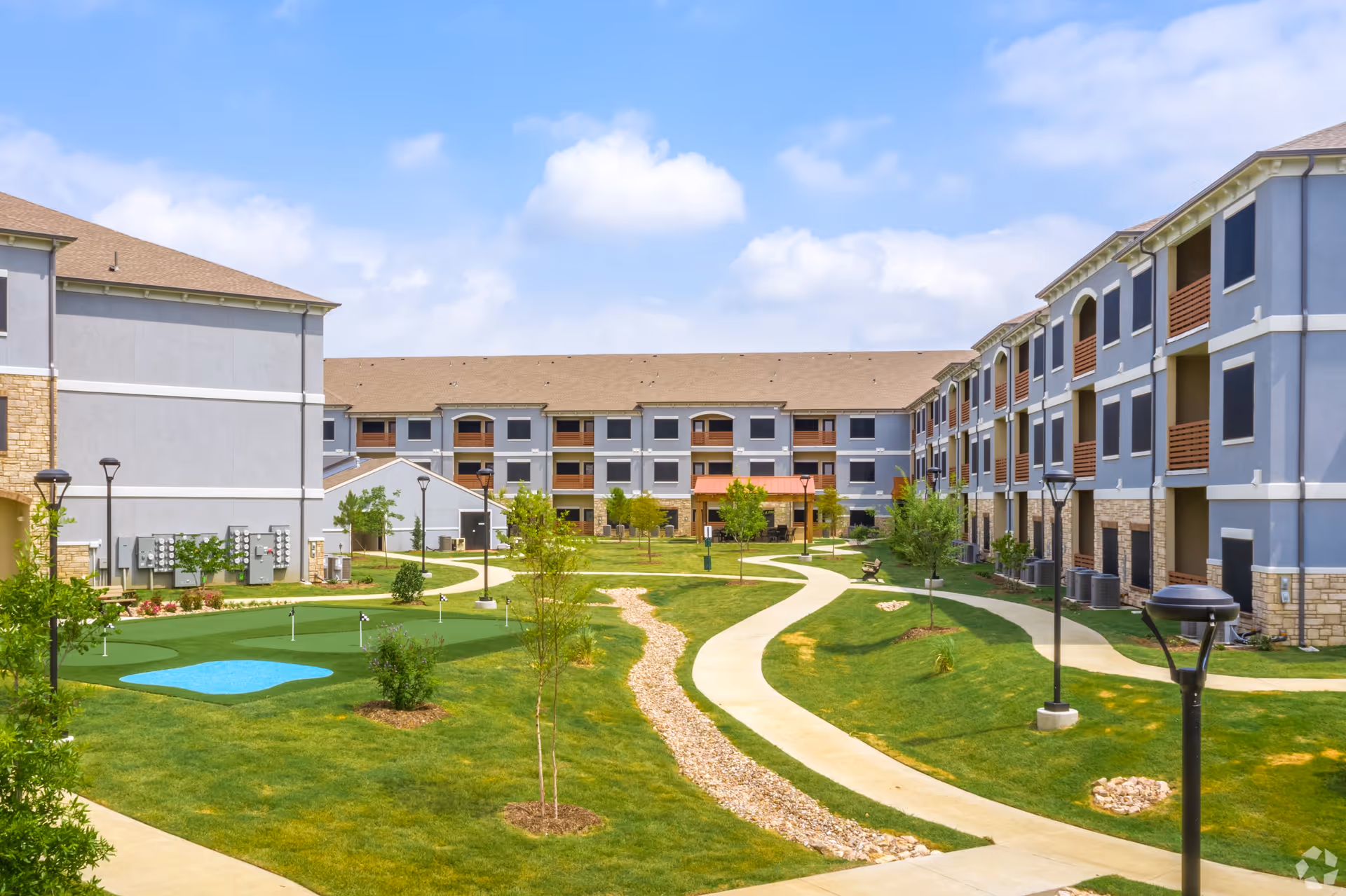 Courtyard of a senior living complex with landscaped lawns, winding walkways, lamp posts, and surrounding three-story residential buildings.
