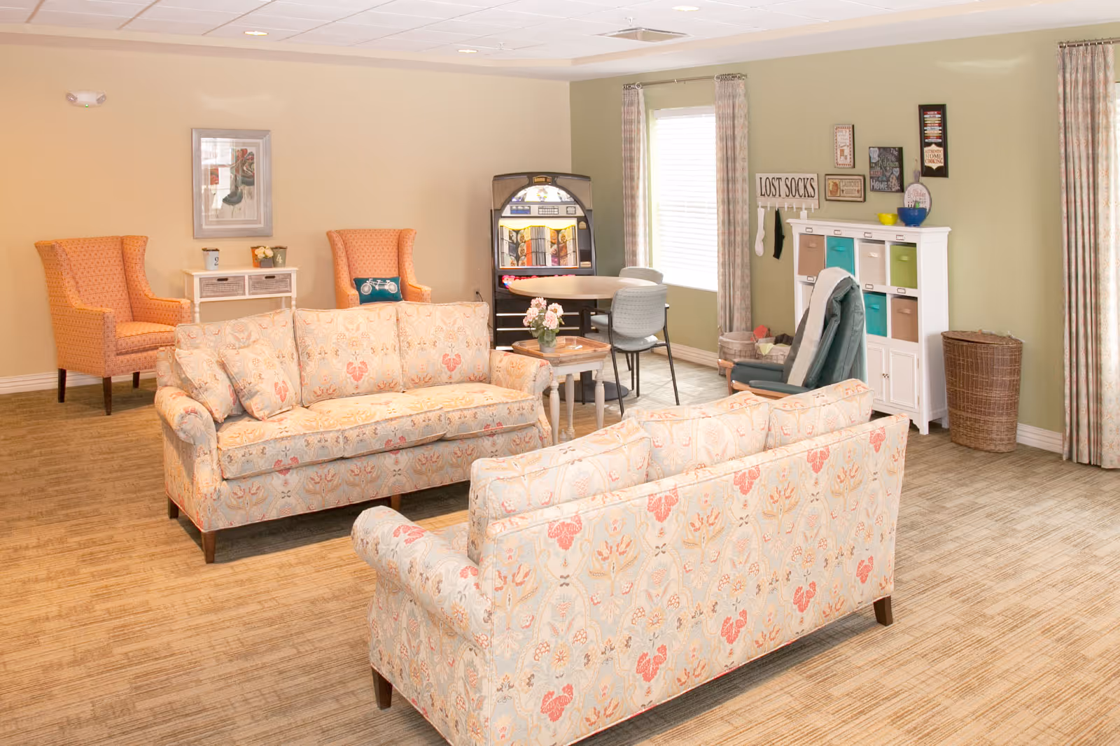 A cozy living room area with two patterned sofas facing each other, two orange armchairs, a small table with flowers, a round table with chairs near a window, a white storage unit with colorful bins, and a sign that reads 'LOST SOCKS' on the wall.