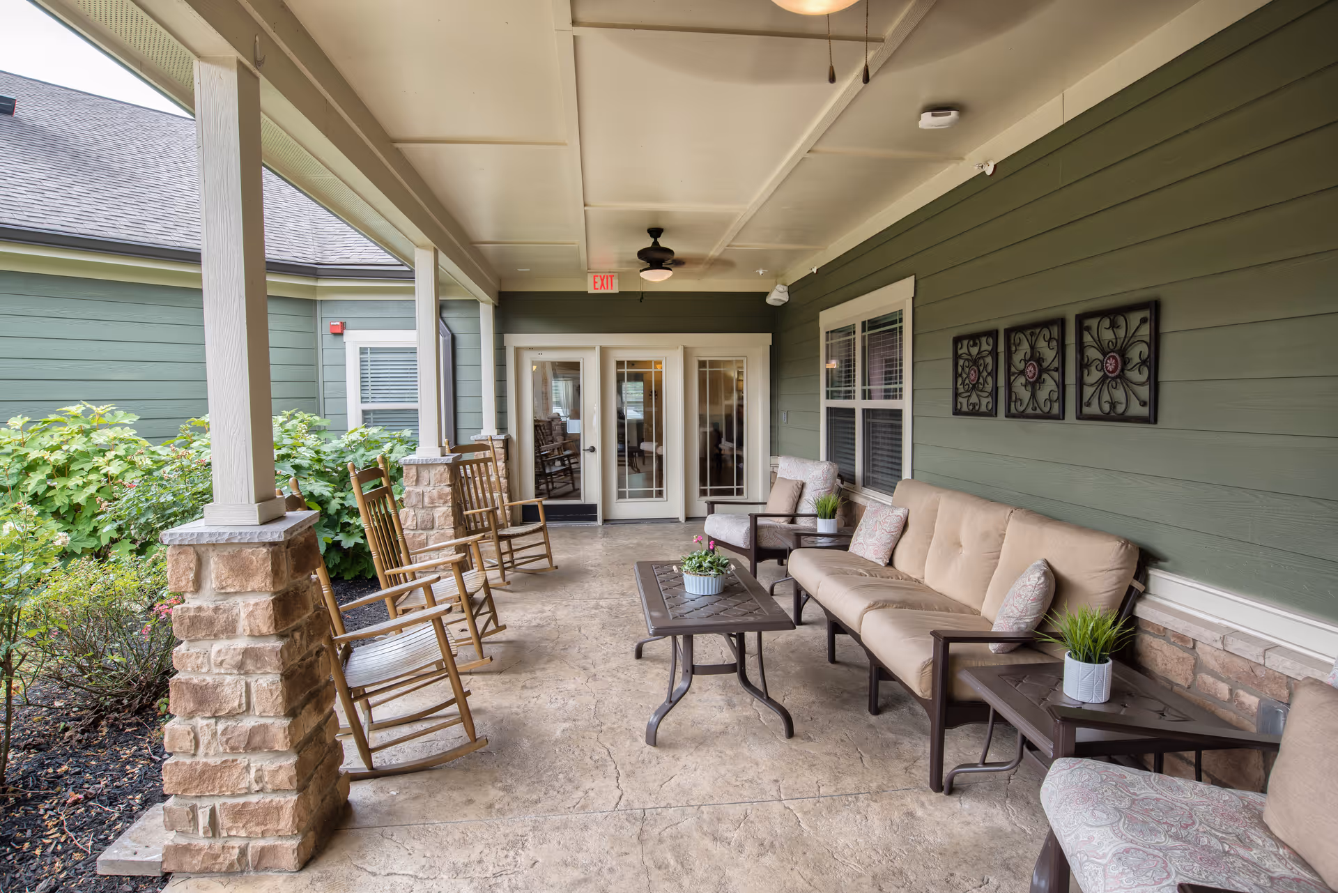 Covered outdoor patio area with cushioned seating including a sofa, armchairs, and rocking chairs. There are small tables with potted plants, decorative wall art, and a ceiling fan. The patio overlooks a garden with green shrubs and is attached to a building with green siding and stone pillars.