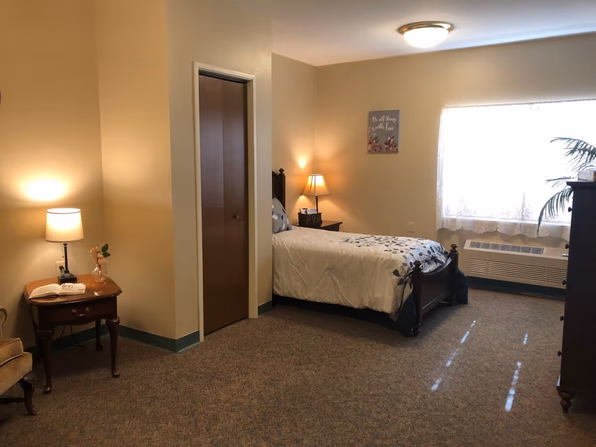 A cozy bedroom with a single bed covered in a white and blue floral comforter. There is a wooden nightstand with a lamp and a basket next to the bed. A window with lace curtains lets in natural light. A wooden dresser is on the right side of the room. On the left, there is a small table with a lamp, a book, and a flower vase. The walls are painted beige, and the floor is carpeted.