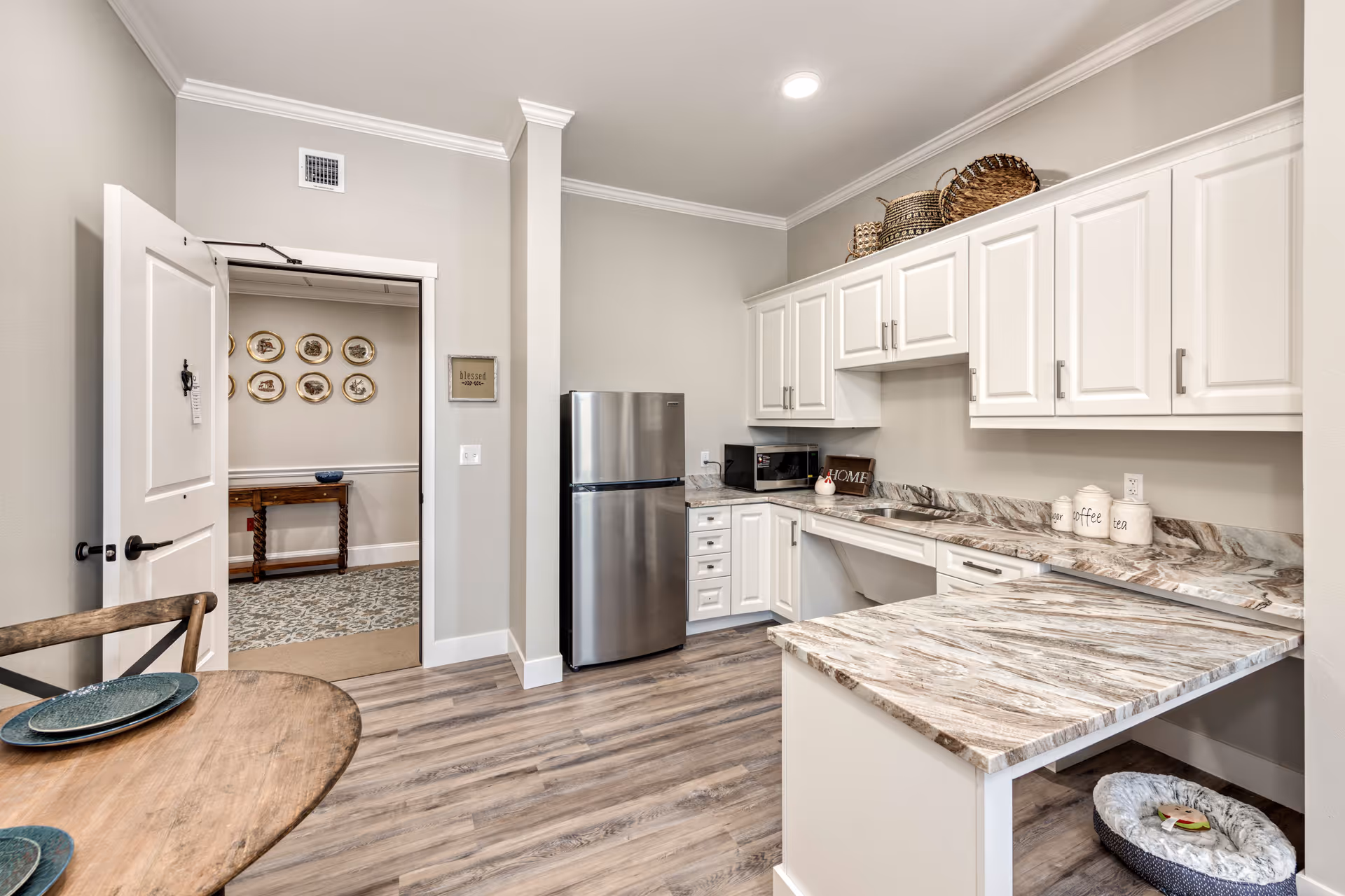 A modern kitchen with white cabinets, marble-patterned countertops, a stainless steel refrigerator, a microwave, and a small round wooden table with two plates. The kitchen has wood flooring and neutral-colored walls. There is an open door leading to another room with decorative plates on the wall and a wooden table.