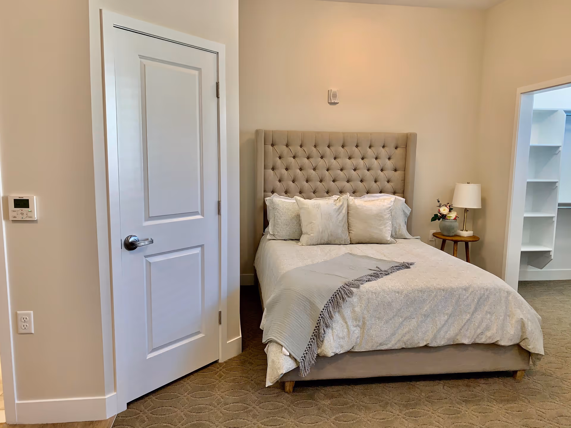 A neatly made bed with a tufted headboard in a softly lit bedroom. The bed has multiple pillows and a light-colored blanket with a fringed throw. To the right of the bed is a small round wooden side table with a lamp and a small flower arrangement. There is a white door on the left side of the image and an open doorway on the right leading to a walk-in closet with shelves.