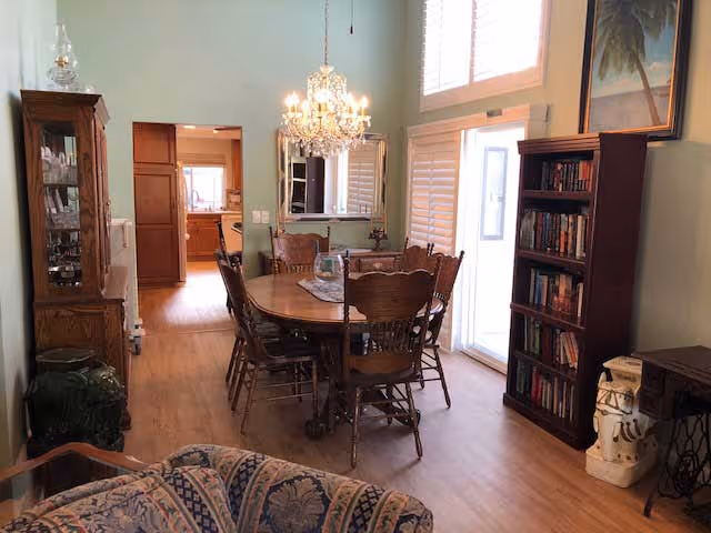 A dining room with a wooden dining table and six chairs under a chandelier. There is a bookshelf filled with books on the right side near a glass door with white shutters. A mirror hangs on the wall above a small table with a lamp. The room has wooden flooring and light green walls. In the background, a kitchen area is visible through an open doorway.