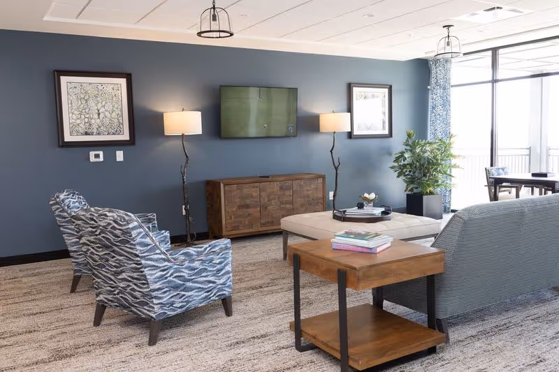 Furnished common living area with upholstered chairs and sofa facing a wall-mounted TV, ottoman, side table, and large windows.