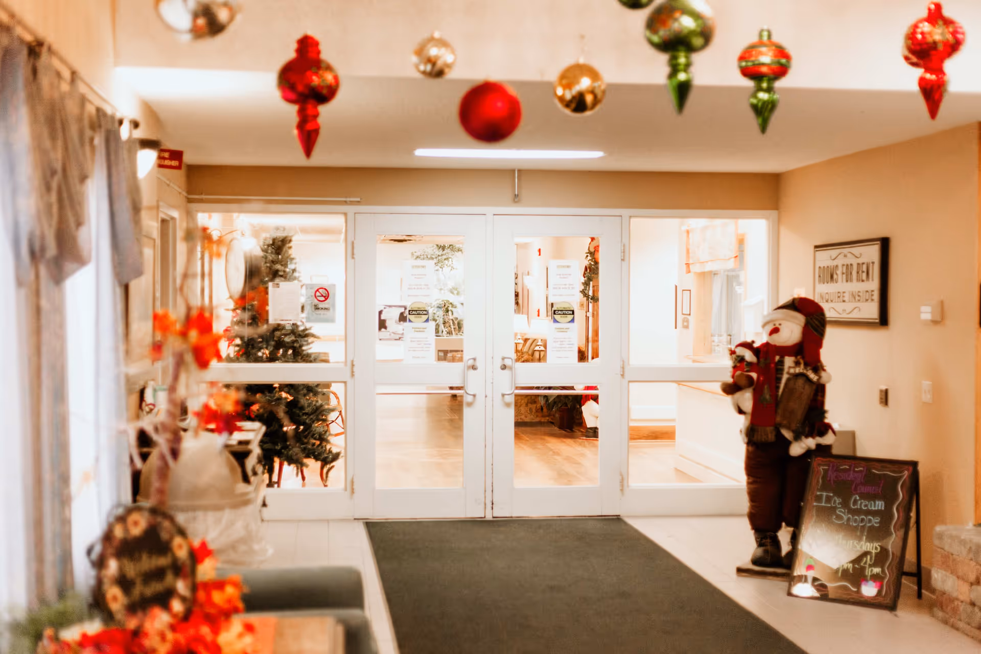 Interior lobby entrance of a senior living community decorated for Christmas with hanging ornaments, a snowman figure, and double glass doors.