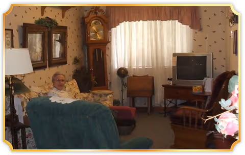 Cozy living room with a person seated in an armchair, a grandfather clock, a TV on a stand, and floral wallpaper and decor.