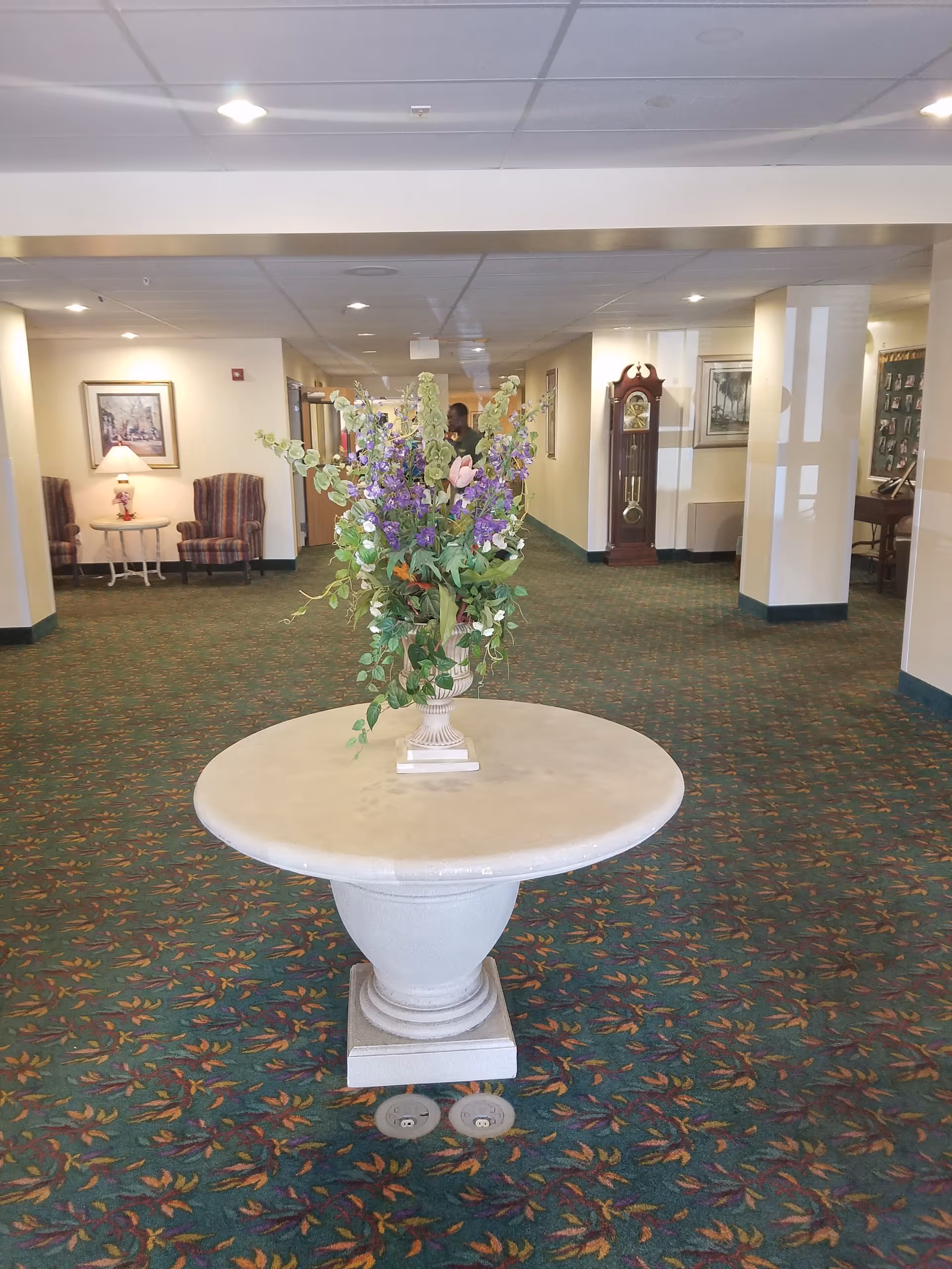 Interior view of a senior living facility hallway with a round table in the center holding a large floral arrangement. The carpet has a colorful leaf pattern. In the background, there are two armchairs with a small table and lamp between them on the left, a grandfather clock on the right, and framed artwork on the walls.