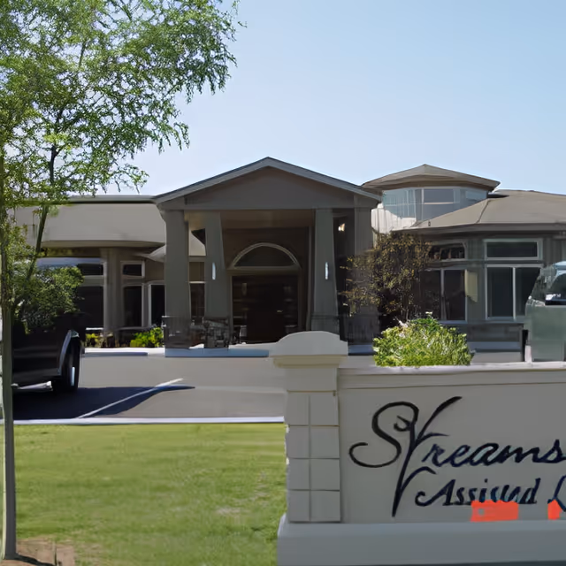 Front exterior view of a single-story building with a covered entrance supported by columns, surrounded by greenery and a parking area. A sign in the foreground partially reads 'Streams Assisted L...'.
