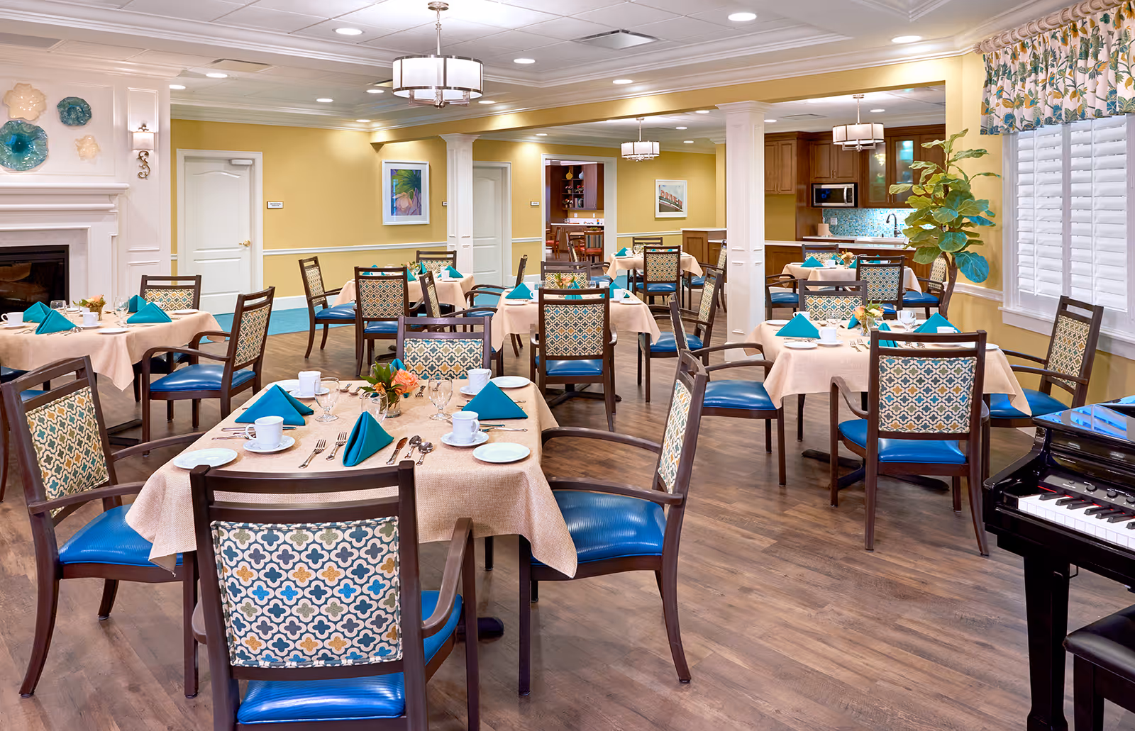 A bright and inviting dining room in an assisted living facility with multiple tables covered in beige tablecloths, each set with white cups, plates, silverware, and teal napkins. The room features wooden chairs with patterned upholstery, hardwood floors, a piano in the corner, and soft yellow walls adorned with framed artwork and decorative plates. Large windows with floral curtains allow natural light to fill the space.