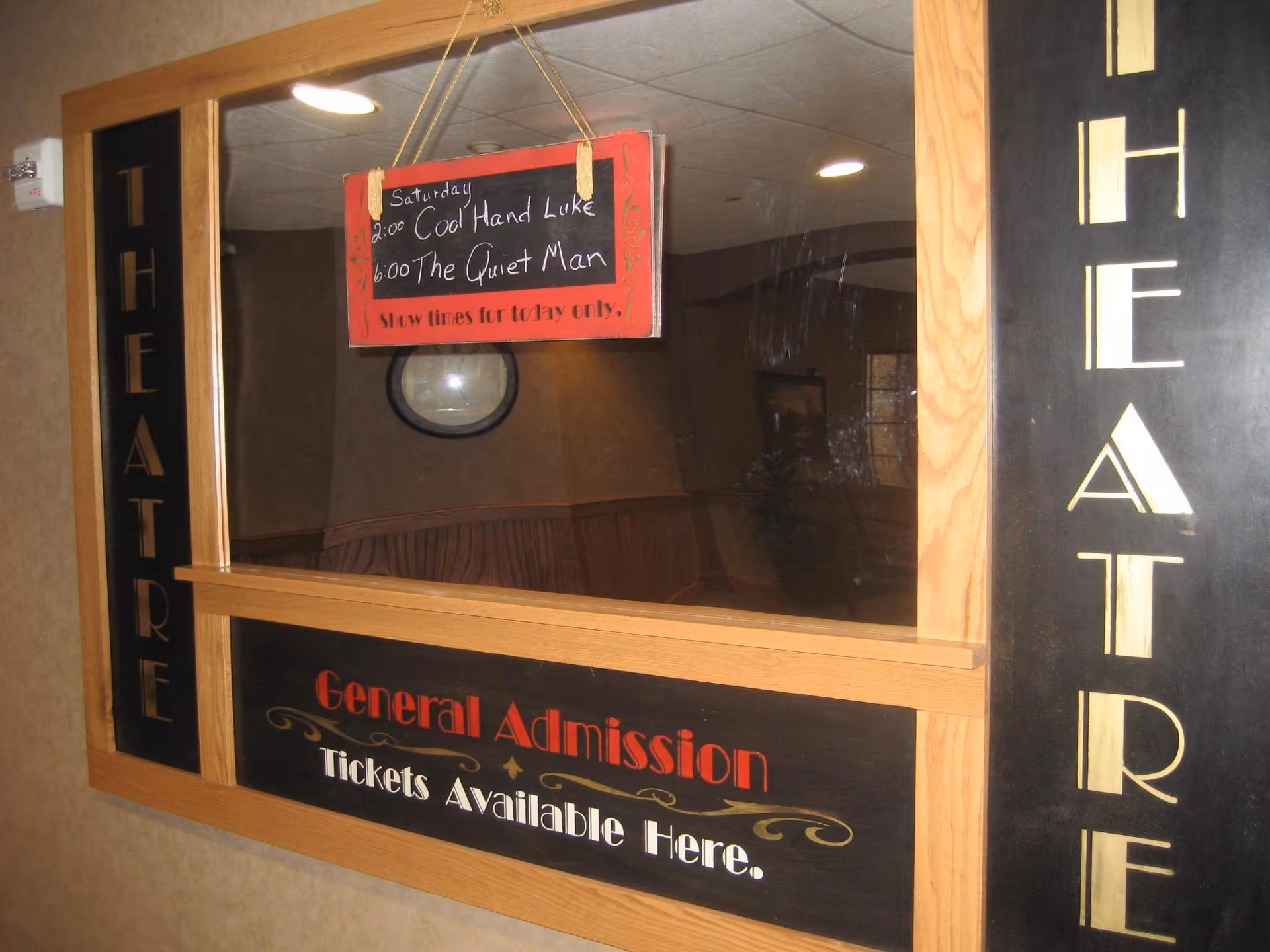 A ticket window for a theatre with wooden framing and black panels on the sides and bottom. The word THEATRE is written vertically on both sides. A small chalkboard sign hanging inside the window lists movie showtimes for Saturday: 2:00 Cool Hand Luke and 6:00 The Quiet Man. Below the window, a sign reads General Admission Tickets Available Here.