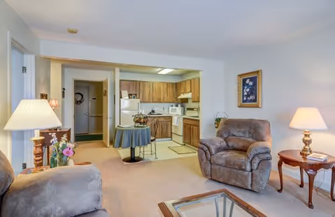 A cozy senior living community apartment featuring a living room with two brown upholstered armchairs, a glass coffee table, and two side tables with lamps. The background shows an open kitchen with wooden cabinets, a white refrigerator, microwave, stove, and a small round dining table with two chairs. The walls are light-colored with a framed floral painting.