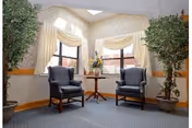 A bright seating area with two upholstered armchairs flanking a small wooden table beneath draped windows, potted plants, and a skylight.