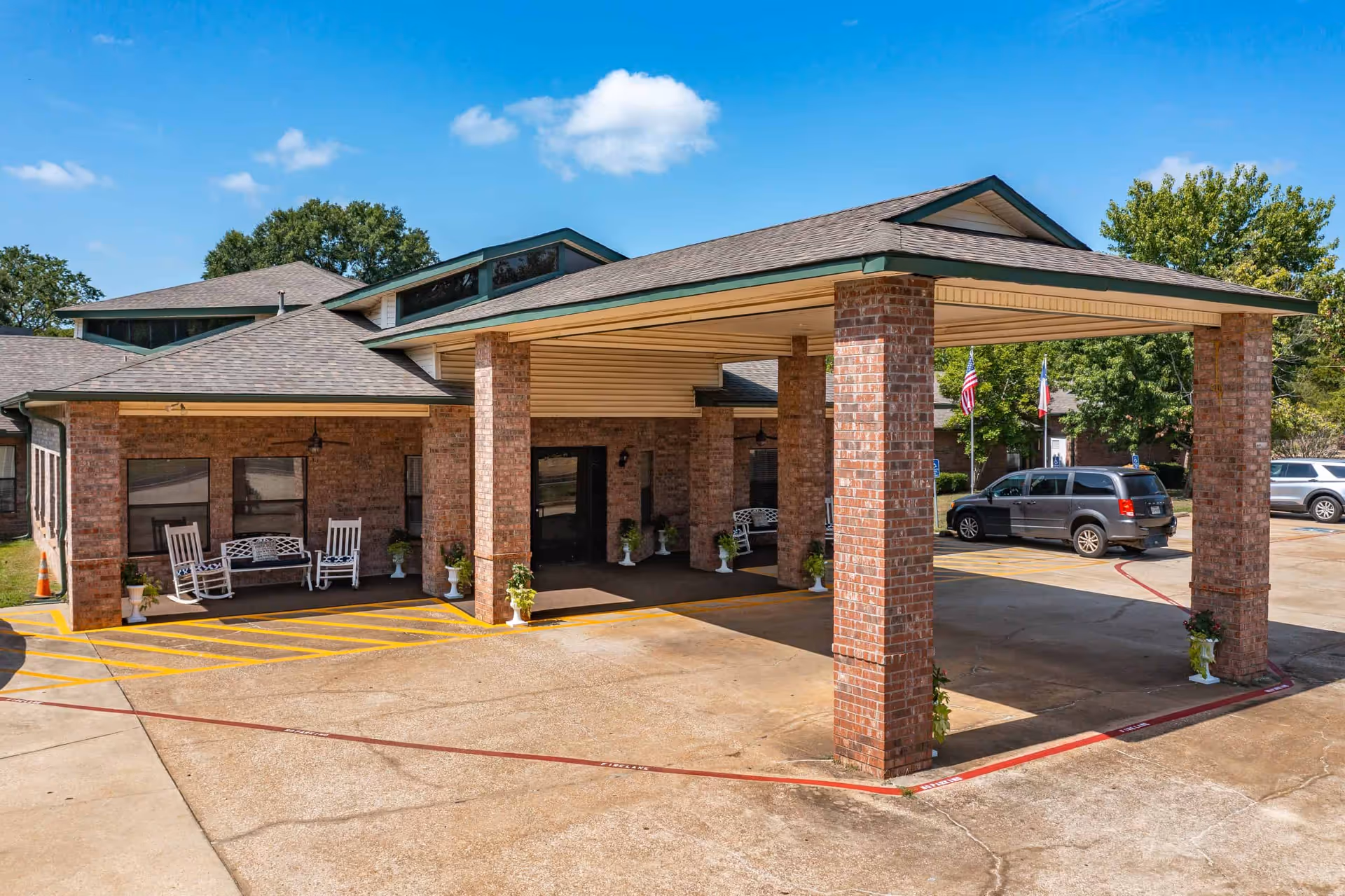 Front entrance of a brick nursing facility showing a covered drive-through canopy with brick columns, rocking chairs on a porch, potted plants, and parked cars.