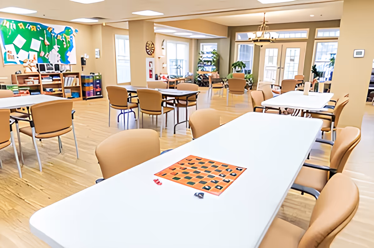 A bright and spacious common area with multiple tables and chairs arranged for group activities or socializing. A checkerboard game is set up on the closest table. The room features large windows letting in natural light, plants, and a bulletin board with colorful papers and decorations on the wall.