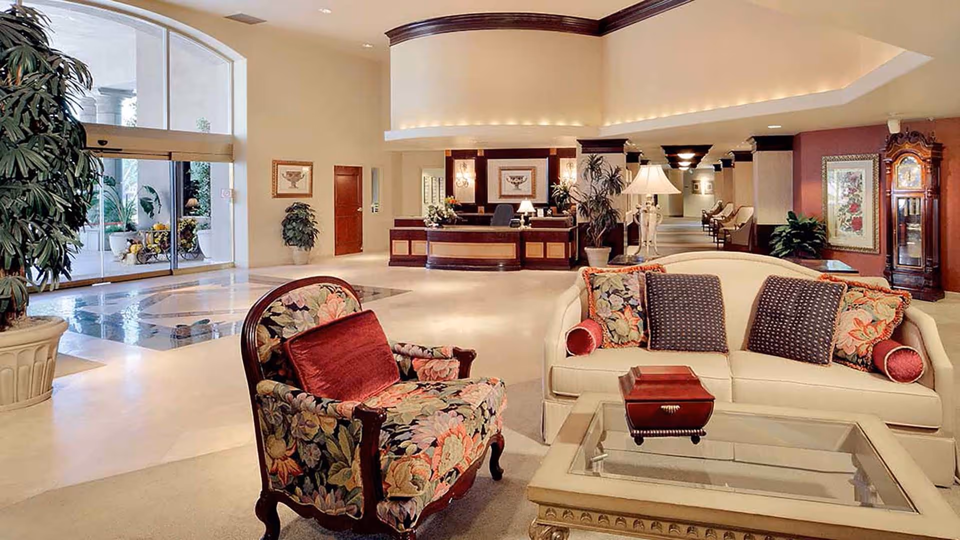 Spacious senior living facility lobby with upholstered sofa and floral armchair, glass coffee table, reception desk and large potted plants.